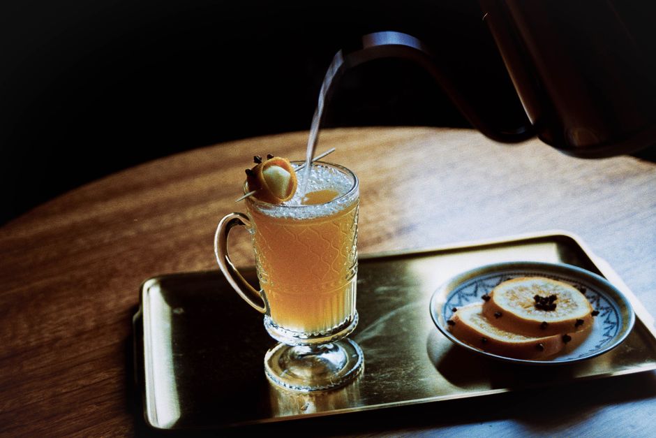 Hot water being poured into a glass mug of hot toddy, with clove-studded citrus on the side